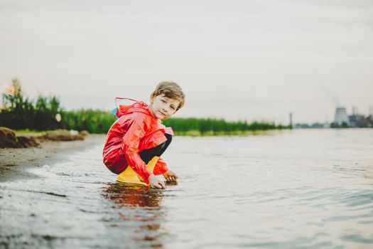 Boy in a red raincoat and yellow rubber boots playing with water at the beach. School kid in a waterproof coat touching water at sea. Child having fun with waves at the shore.