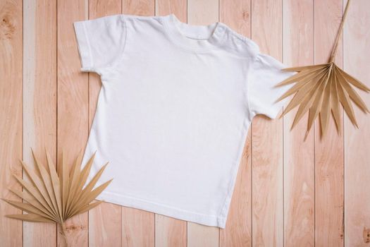 White baby t-shirt top view. Mock-up for logo, text or design on wooden background. Flat lay child clothes with palm leaves