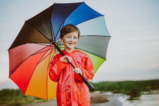 Portrait of a smiling school boy with rainbow umbrella in the park. Kid holds colourful umbrella on his shoulder. Cheerful child in a red raincoat holding multicolor umbrella outdoors.