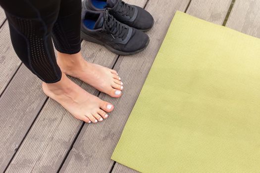 Close-up of bare feet on a wooden floor near a green sports mat, there are black sneakers nearby, in summer, near a pond in the park. Top view. Mock up