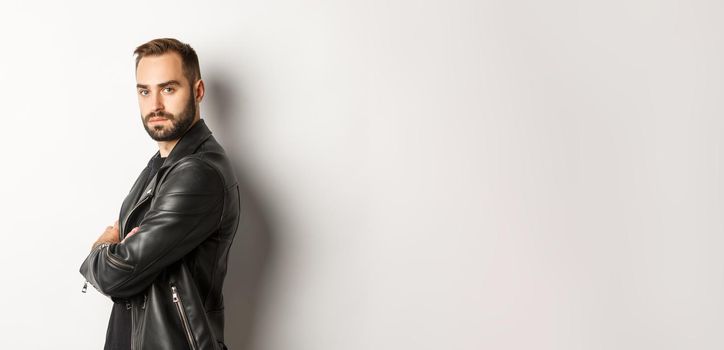 Profile of confident and handsome bearded man in black leather jacket, turn face at camera and looking serious, white background.