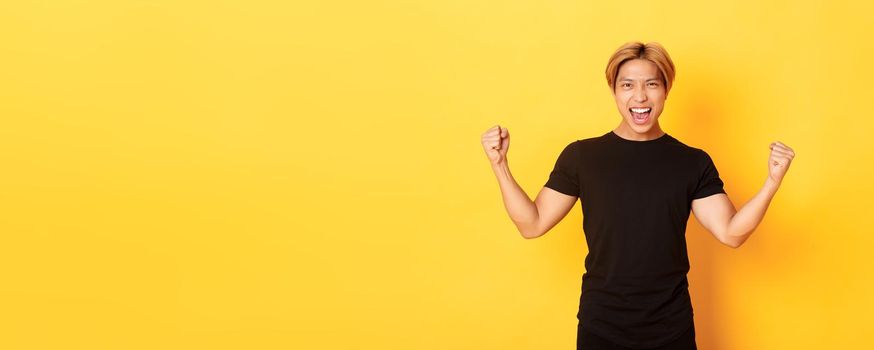 Portrait of happy attractive asian man rejoicing, raising hands up and shouting yes, standing pleased over yellow background.