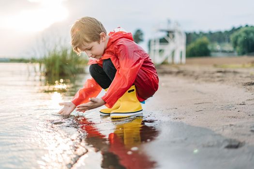 Boy in a red raincoat and yellow rubber boots playing with water at the beach. School kid in a waterproof coat touching water at sea. Child having fun with waves at the shore.