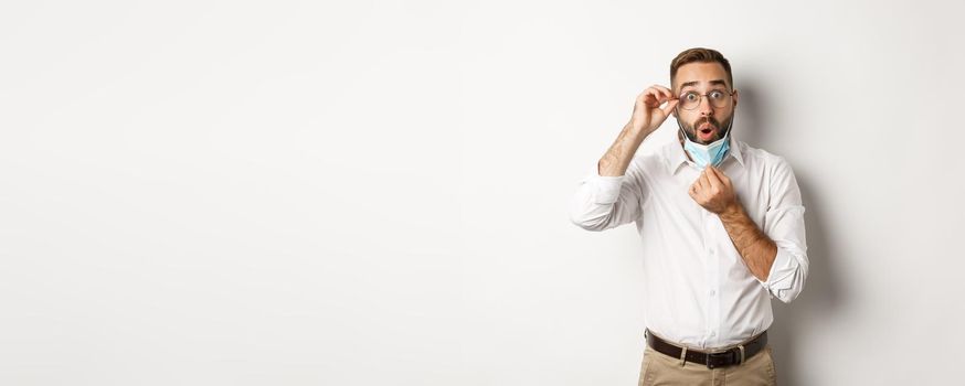 Covid-19, social distancing and quarantine concept. Impressed business man take off medical mask, looking surprised, standing over white background.