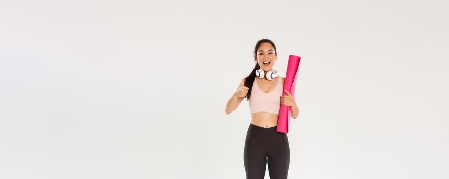 Full length of cheerful satisfied, slim asian girl with fitness rubber mat and headphones, female athlete showing thumbs-up, recommend gym or yoga classes, looking pleased after good workout.