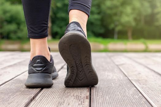 Close-up of women's legs in black sneakers and black leggings walking on a wooden deck in a park, in summer. Copy space