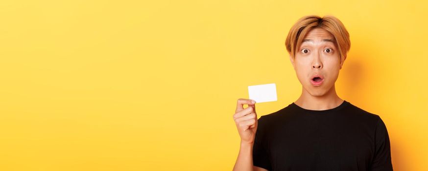 Close-up of surprised gasping asian guy with fair hair, looking amazed and startled as showing credit card, standing yellow background.