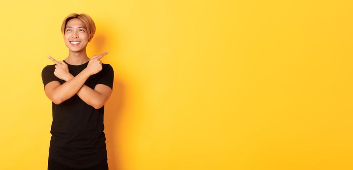 Portrait of smiling pleased asian guy, looking left and making decision, pointing sideways, yellow background.