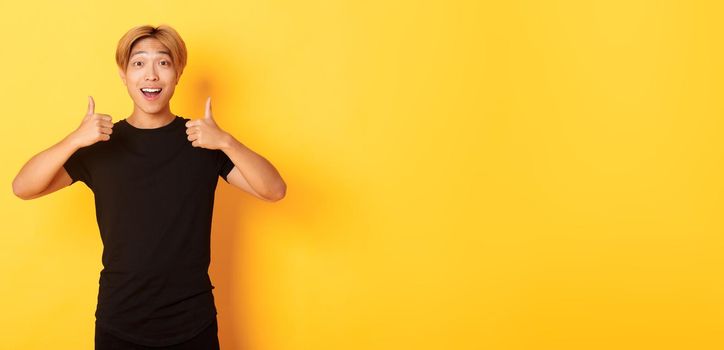 Portrait of amazed and pleased asian guy in black outfit, showing thumbs-up in approval, standing excited over yellow background.