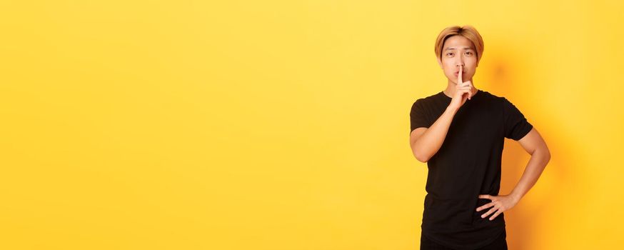 Portrait of attractive blond asian man shushing, press finger to lips and hush, yellow background.