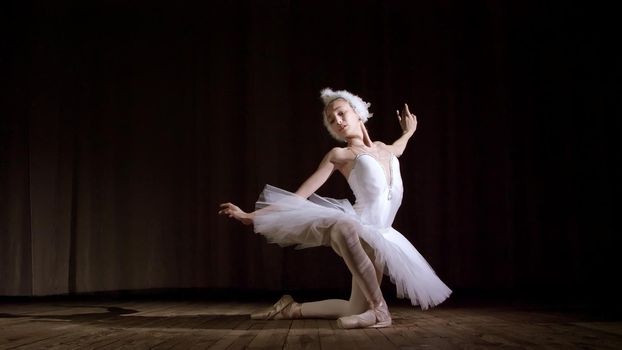in rays of spotlight, on the stage of the old theater hall. Young ballerina in suit of white swan and pointe shoes, dances elegantly certain ballet motion, part de bras. High quality photo