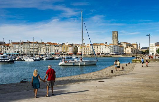 View of the old harbour of the French city of La Rochelle with tourists strolling and a ship entering the harbour, with blue sky and light white clouds.