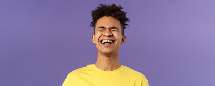 Close-up portrait of happy carefree young guy laughing loud, chuckling over hilarious joke, bending backwards and close eyes while giggle over funny movie, purple background.