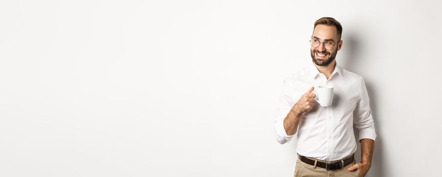 Successful business man drinking coffee, looking sideways with satisfied smile, standing over white background.