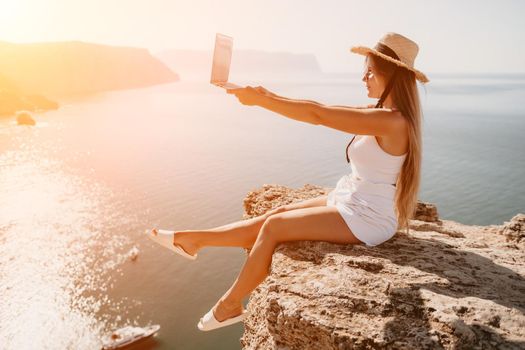 Successful business woman in yellow hat working on laptop by the sea. Pretty lady typing on computer at summer day outdoors. Freelance, travel and holidays concept.