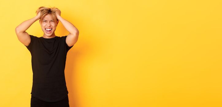 Portrait of pissed-off angry asian man tossing haircut and yelling furious, standing over yellow background.