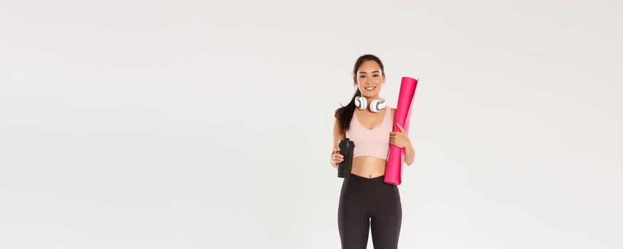 Full length of cute brunette asian fitness girl with headphones, activewear and water bottle, carry pink rubber mat for gym exercises, going for yoga classes, standing over white background.