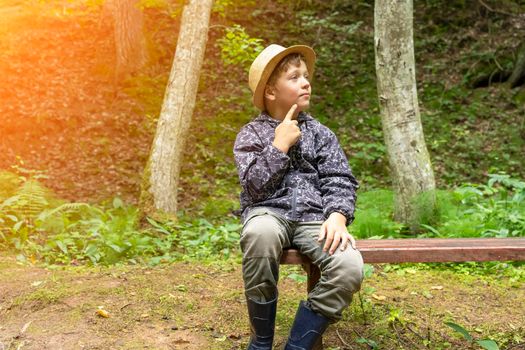funny Caucasian boy in a traveler's hat and windbreaker is sitting on a bench in a nature park near a forest stream and rocks, looking thoughtfully away