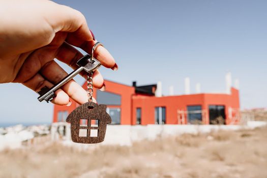 A woman's hand holds a house key against the backdrop of a house under construction. Real estate agent. Buying a house, apartment