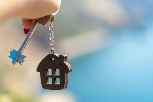 Keys with a keychain in the shape of a house in a female hand against the backdrop of the sea. The concept of buying a house, apartment, real estate, young family, mortgage loan, rental housing