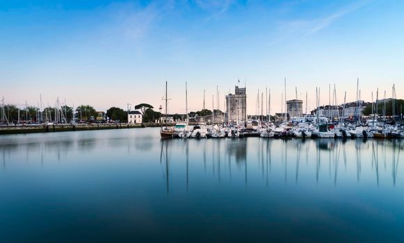 View of the old port of the French city of La Rochelle at sunset with ships at anchor in the harbour and the two towers in the background.