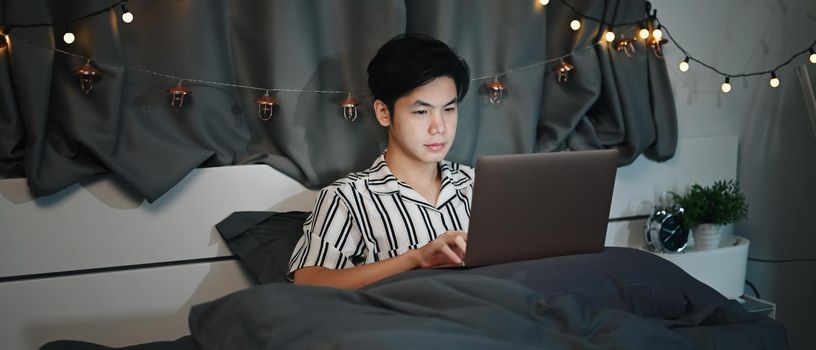 Young man in casual pajamas sitting in bed and working on laptop computer at night.