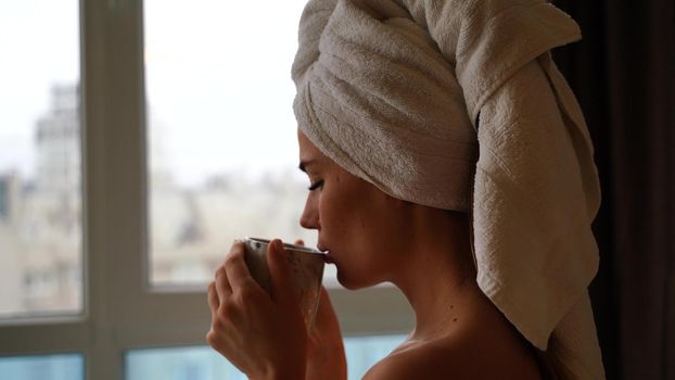 Middle aged woman looks good with bare shoulders in a white towel on her head holds a cup and drinks coffee or tea against the window.