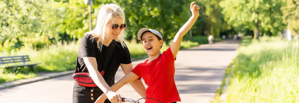 Beautiful and happy young mother teaching her daughter to ride a bicycle. Both smiling, summer park in background