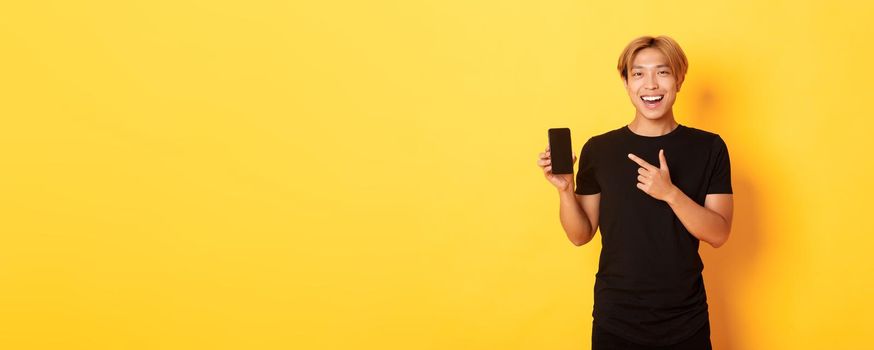 Portrait of satisfied smiling asian guy with blond hair, looking amused and pointing finger at smartphone screen, standing yellow background.