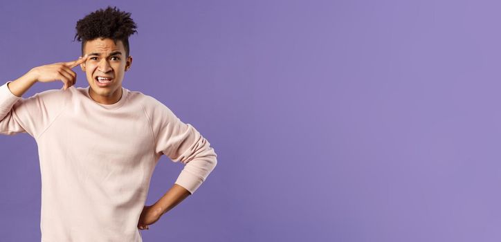 Portrait of angry, annoyed young man scolding someone from being stupid and crazy, rolling index finger over temple staring outraged and irritated camera, standing bothered purple background.