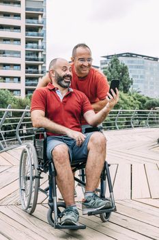 disabled man in wheelchair watching the phone and laughing with a friend during a walk, concept of friendship and integration of people with disabilities and reduced mobility problems, vertical photo