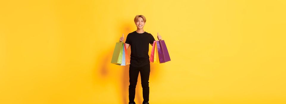 Full-length of attractive korean guy in black clothes, showing thumbs-up, holding shopping bags, yellow background.