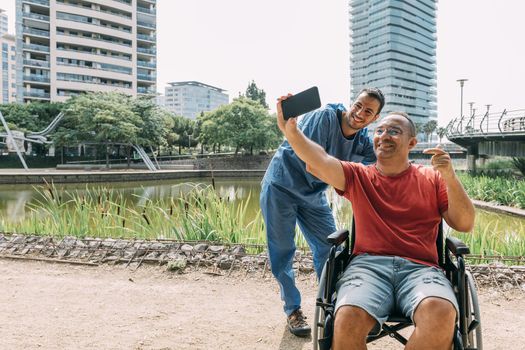 disabled man in a wheelchair taking a selfie with his caretaker during a walk, concept of medical care and rehabilitation of people with disabilities and reduced mobility problems