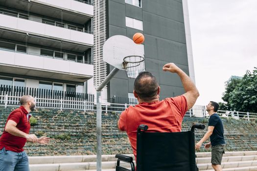 joyful disabled man in wheelchair throwing the ball to basket with two friends, concept of adaptive sports and physical activity, rehabilitation for people with physical disabilities