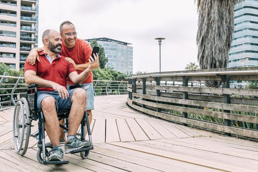 disabled man in wheelchair making a selfie with his phone with a friend during a walk, concept of friendship and integration of people with disabilities and reduced mobility problems