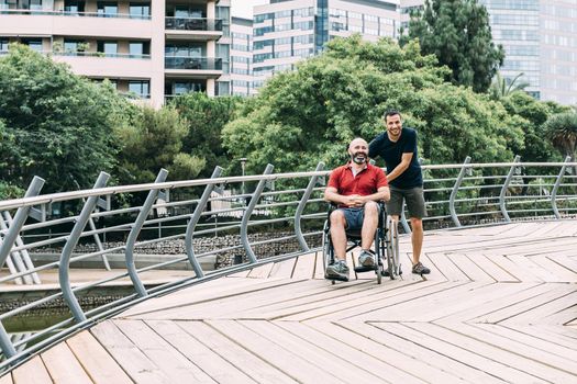 disabled man in wheelchair walking and laughing in the park with a friend, concept of friendship and integration of people with disabilities and reduced mobility problems
