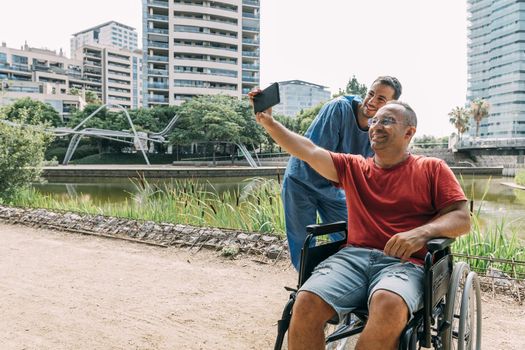 disabled man in a wheelchair taking a photo with his caretaker during a walk, concept of medical care and rehabilitation of people with disabilities and reduced mobility problems