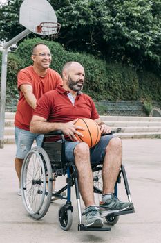 man pushing the wheelchair to play basketball with his handicapped friend, concept of adaptive sports and physical activity, rehabilitation for people with physical disabilities, vertical photo