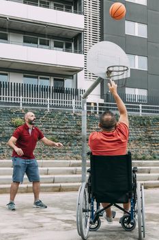 joyful handicapped man in wheelchair throwing the ball to basket with a friend, concept of adaptive sports and physical activity, rehabilitation for people with physical disabilities, vertical photo