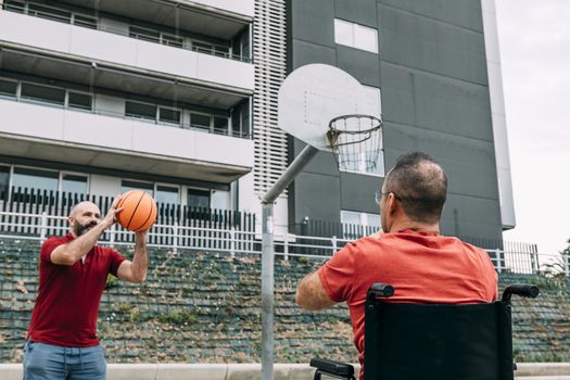 joyful disabled man in wheelchair playing basketball with a friend with a ball, concept of adaptive sports and physical activity, rehabilitation for people with physical disabilities