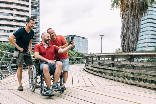 disabled man in wheelchair taking a selfie with his phone with two friends during a walk, concept of friendship and integration of people with disabilities and reduced mobility problems