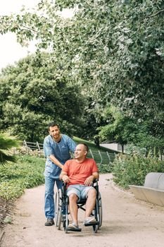 disabled man in wheelchair talking during a walk with his caretaker at park, concept of medical care and rehabilitation of people with disabilities and reduced mobility problems, vertical photo