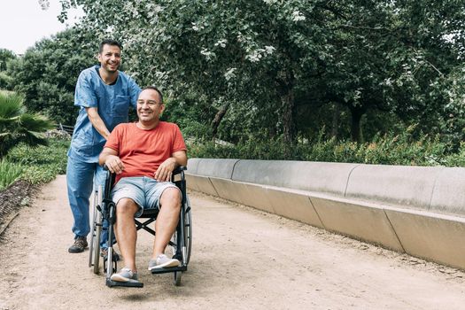 disabled man in wheelchair laughing during a walk with his nurse at park, concept of medical care and rehabilitation of people with disabilities and reduced mobility problems