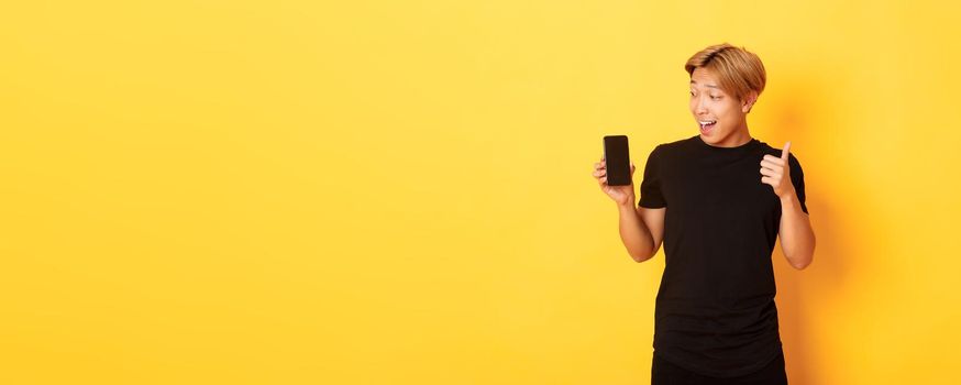 Portrait of satisfied asian guy looking at smartphone screen and showing thumbs-up in approval, standing yellow background.
