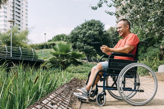 handicapped man in wheelchair having fun while working using a tablet computer at park, concept of technological and occupational integration of people with disabilities and reduced mobility problems