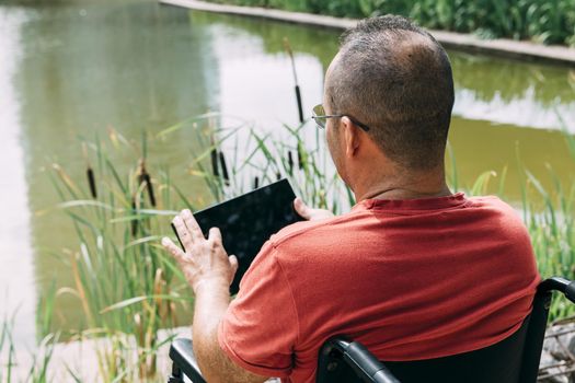 handicapped man in wheelchair having fun while resting using a tablet computer at park, concept of technological and occupational integration of people with disabilities and reduced mobility problems
