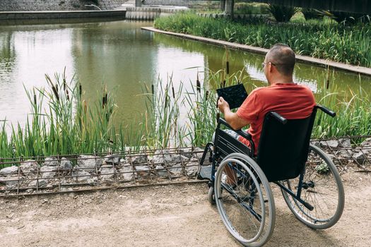 disabled man in wheelchair having fun while resting using a tablet computer at park, concept of technological and occupational integration of people with disabilities and reduced mobility problems