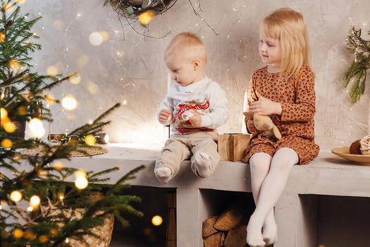 Little brother and sister play on Christmas eve in a beautiful house decorated for the New Year holidays. Children are playing with a Christmas gift. Scandinavian-style interior with live fir trees and a wooden staircase.