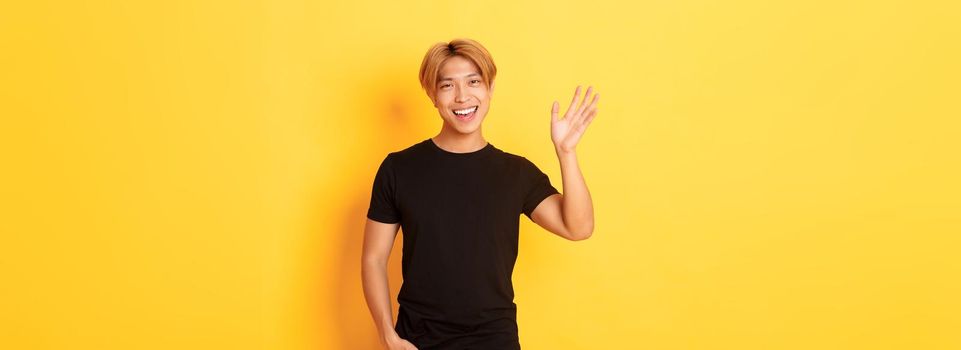 Portrait of handsome friendly asian guy in black outfit, waving hand to say hello and smiling, greeting someone, standing yellow background.