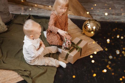 Little brother and sister play on Christmas eve in a beautiful house decorated for the New Year holidays. Children are playing with a Christmas gift. Scandinavian-style interior with live fir trees and a wooden staircase.